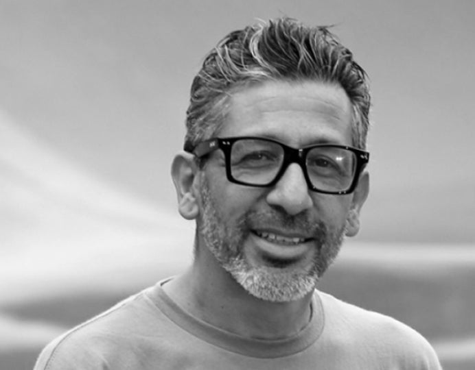 A man with short, wavy hair and a beard smiles at the camera. He is wearing dark, thick-rimmed glasses and a casual shirt. The background is softly blurred and monochrome.