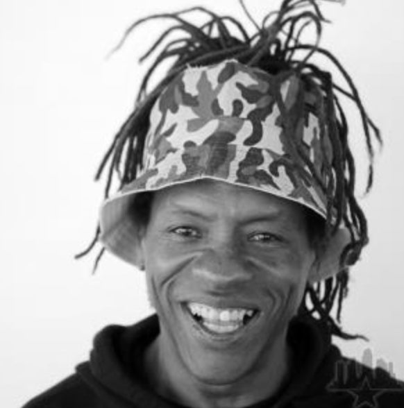 A smiling person wearing a camouflage-patterned bucket hat with dreadlocks sticking out, dressed in a dark hoodie, pictured against a plain white background.