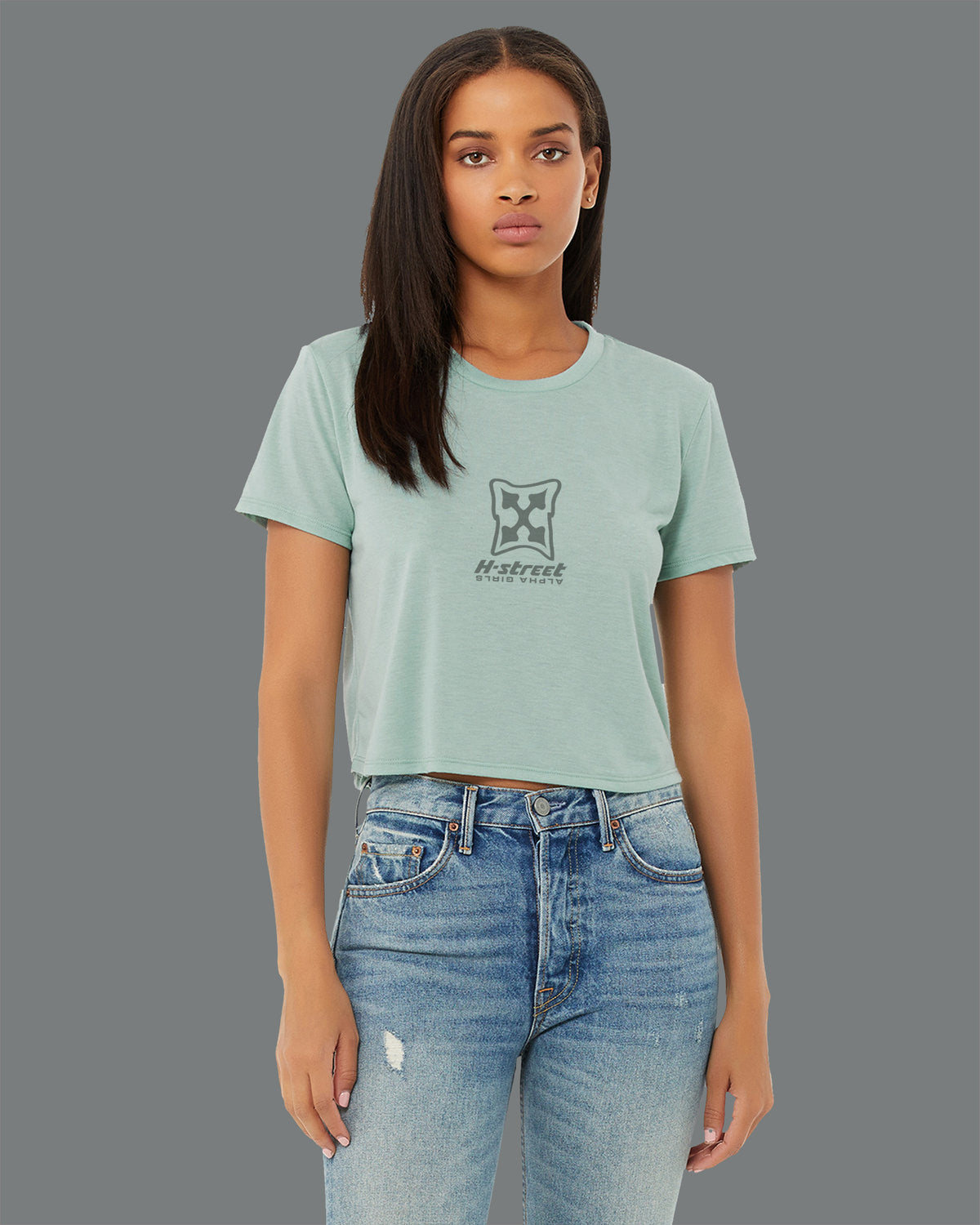 A woman with straight, dark hair wears an MH Printing ALPHA GIRL MARK LOGO Flowy Crop T-Shirt in light green and light blue ripped jeans, standing against a plain gray background and channeling an Alpha Girl vibe as she looks at the camera.
