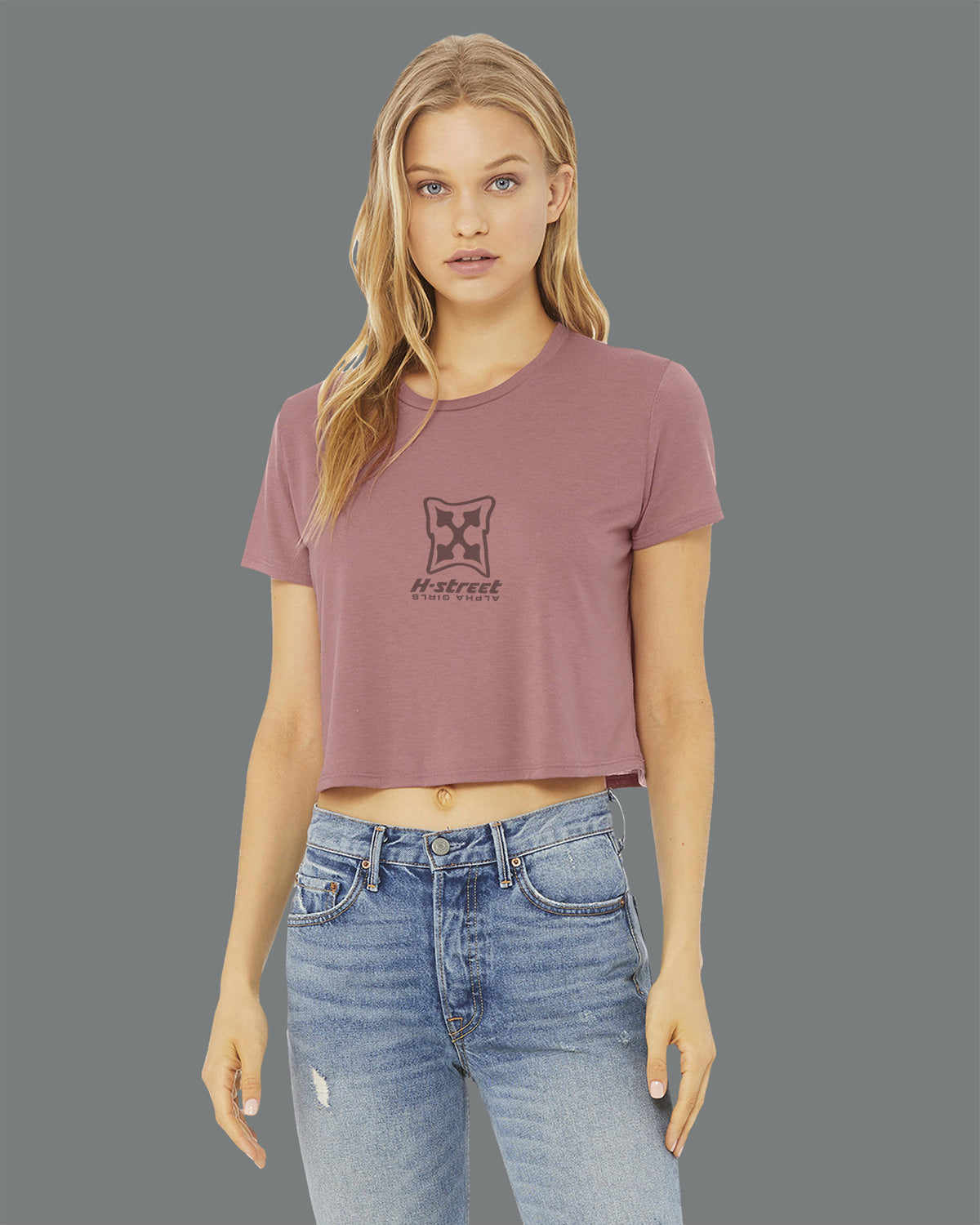 A woman with long blonde hair wears the MH Printing ALPHA GIRL MARK LOGO flowy crop t-shirt, featuring a geometric graphic and text, styled with light blue jeans against a plain gray background.