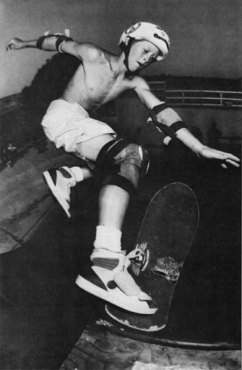A young skateboarder wearing a helmet and protective pads performs a trick in mid-air on a ramp, with one hand outstretched and focused expression, in a black and white photo.