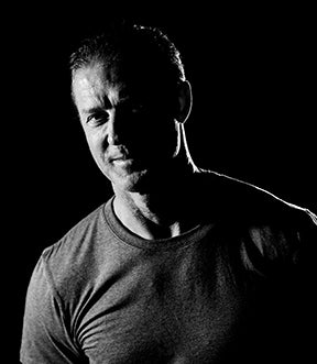 A man in a fitted t-shirt stands against a black background, partially lit from the side, creating dramatic shadows on his face and body.