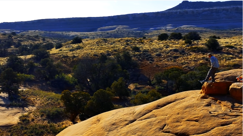 A person stands on a sunlit orange rock formation overlooking a wide desert landscape with trees, rolling hills, and distant mesas under a pale sky.