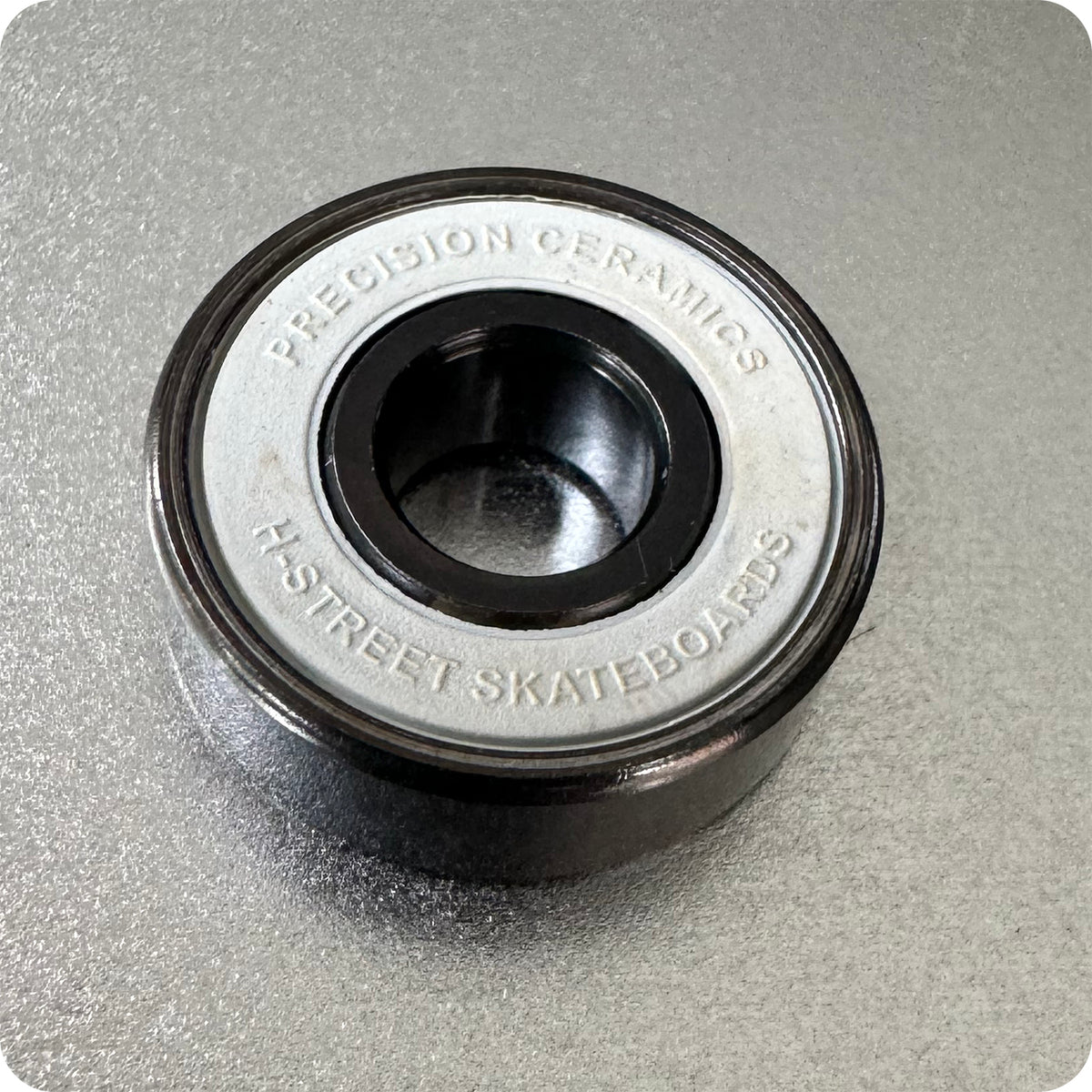 A close-up photo of an Art Function HST FAF Bearings skateboard bearing, with the product name engraved on the metal shield, placed on a textured gray surface and highlighting top-tier ceramic performance.