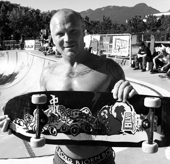 A shirtless man holding a skateboard stands by a skatepark bowl, with mountains and trees in the background. People are sitting and watching nearby. The image is in black and white.