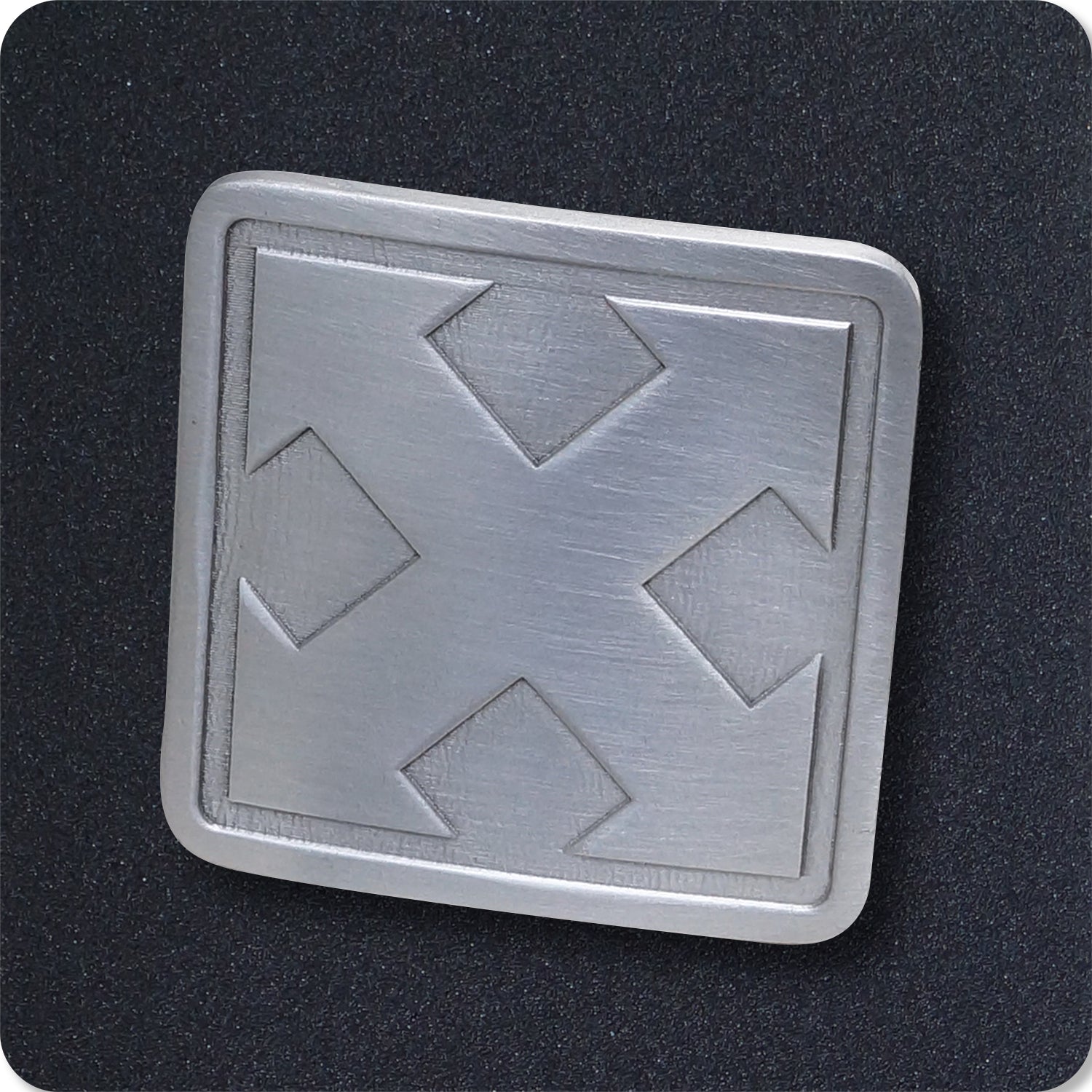 A square metallic badge with four arrows pointing outward from the center, set against a textured black background. The badge has a slightly brushed finish and rounded corners.
