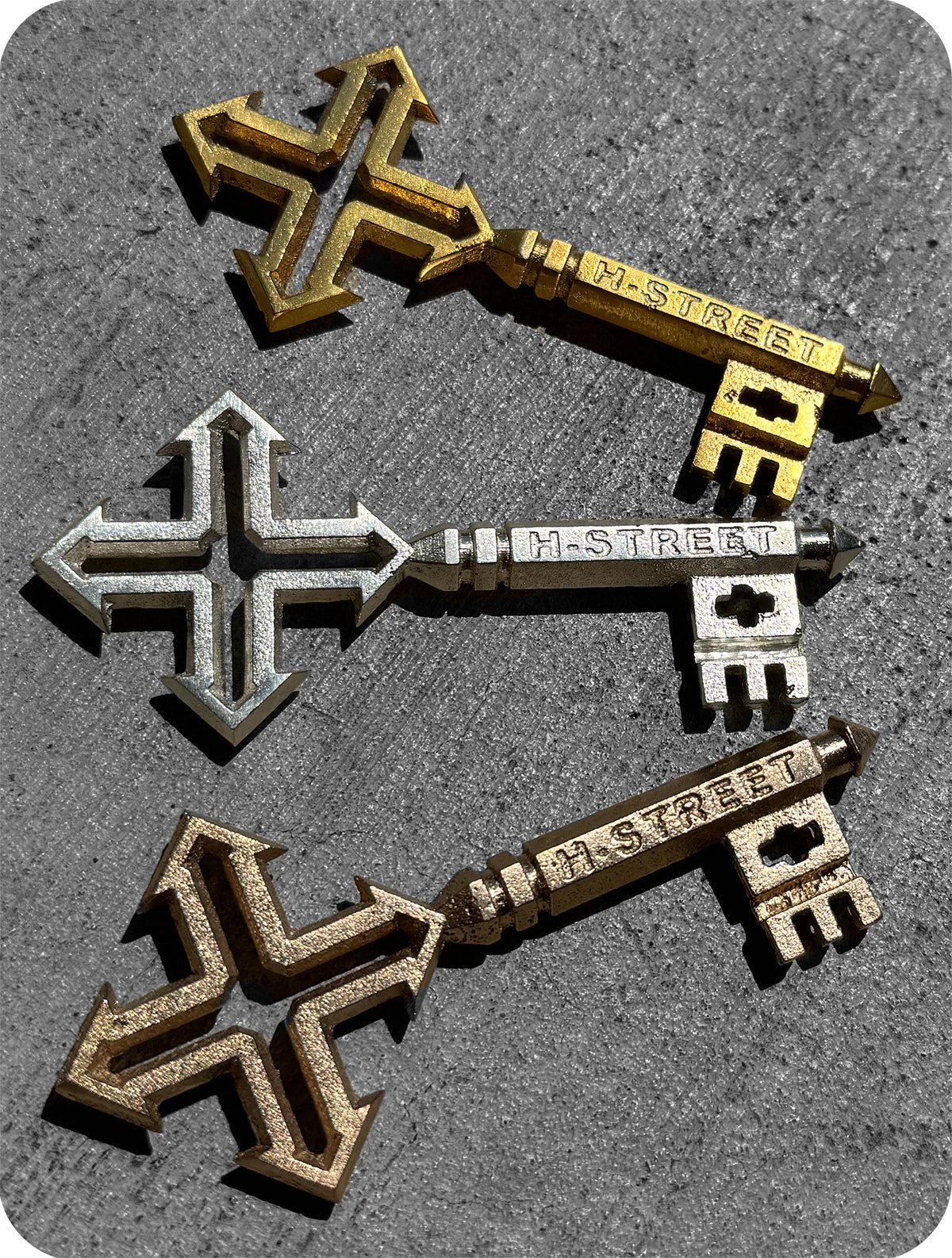 Three metal keys—gold, silver, and bronze—from Perfect Skateboarding’s STREET KEY OG A SERIES are arranged diagonally on a textured gray surface. Each has a stylized cross-shaped head and unique, blocky teeth.