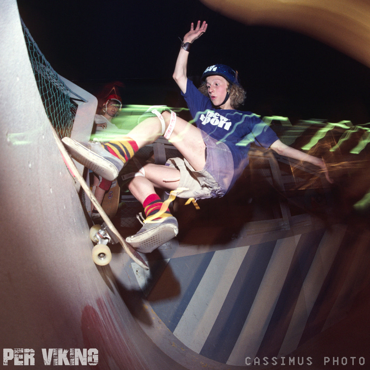 Wearing a helmet, knee pads, and striped socks, a skateboarder rides up a ramp at night in Vällingby as another watches. Motion blur highlights the action. Text: PER VIKING MEMORIAL SERIES by Woodchuck and CASSIMUS PHOTO.