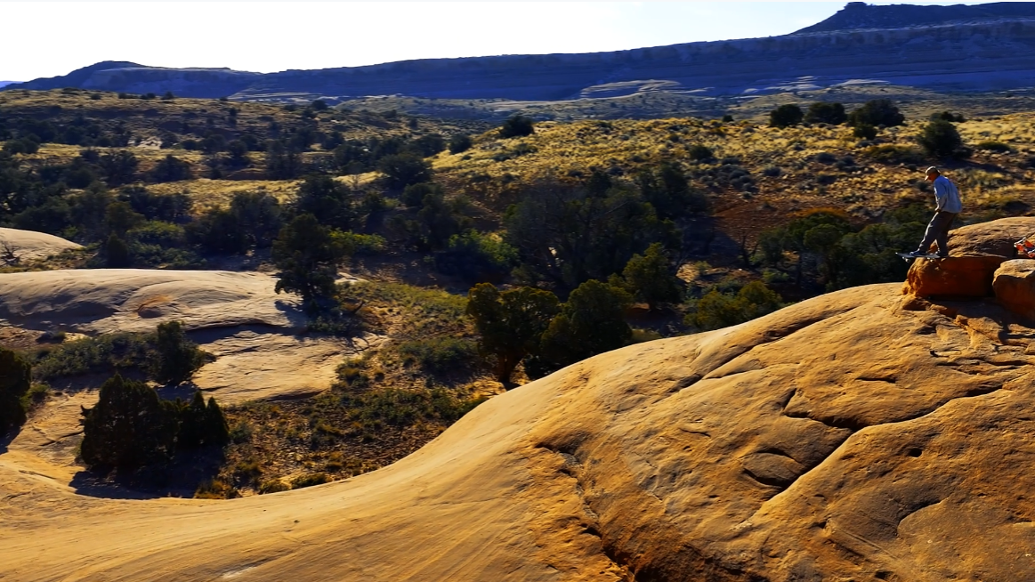 A wide outdoor landscape of sunlit tan rock formations and scrub vegetation under a pale sky, with a small person standing on a rock outcrop in the upper right and distant blue-gray mountains on the horizon.
