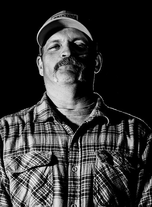 Black and white portrait of a man with a mustache wearing a plaid shirt and a baseball cap, facing forward against a dark background. The lighting creates dramatic shadows on his face and shirt.