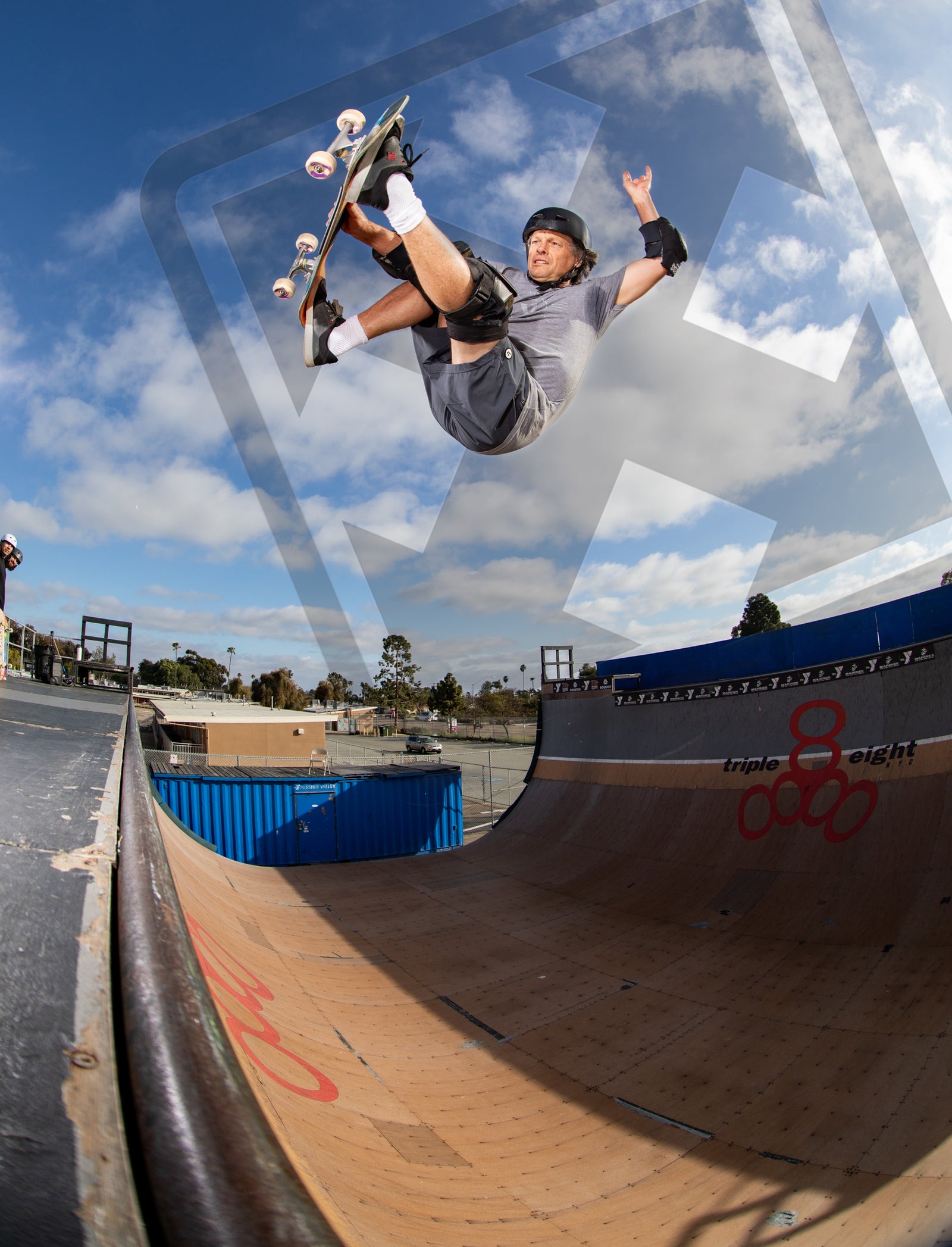A skateboarder wearing a helmet and pads performs an aerial trick above a halfpipe under a partly cloudy sky, with one arm raised and legs bent, at an outdoor skate park.