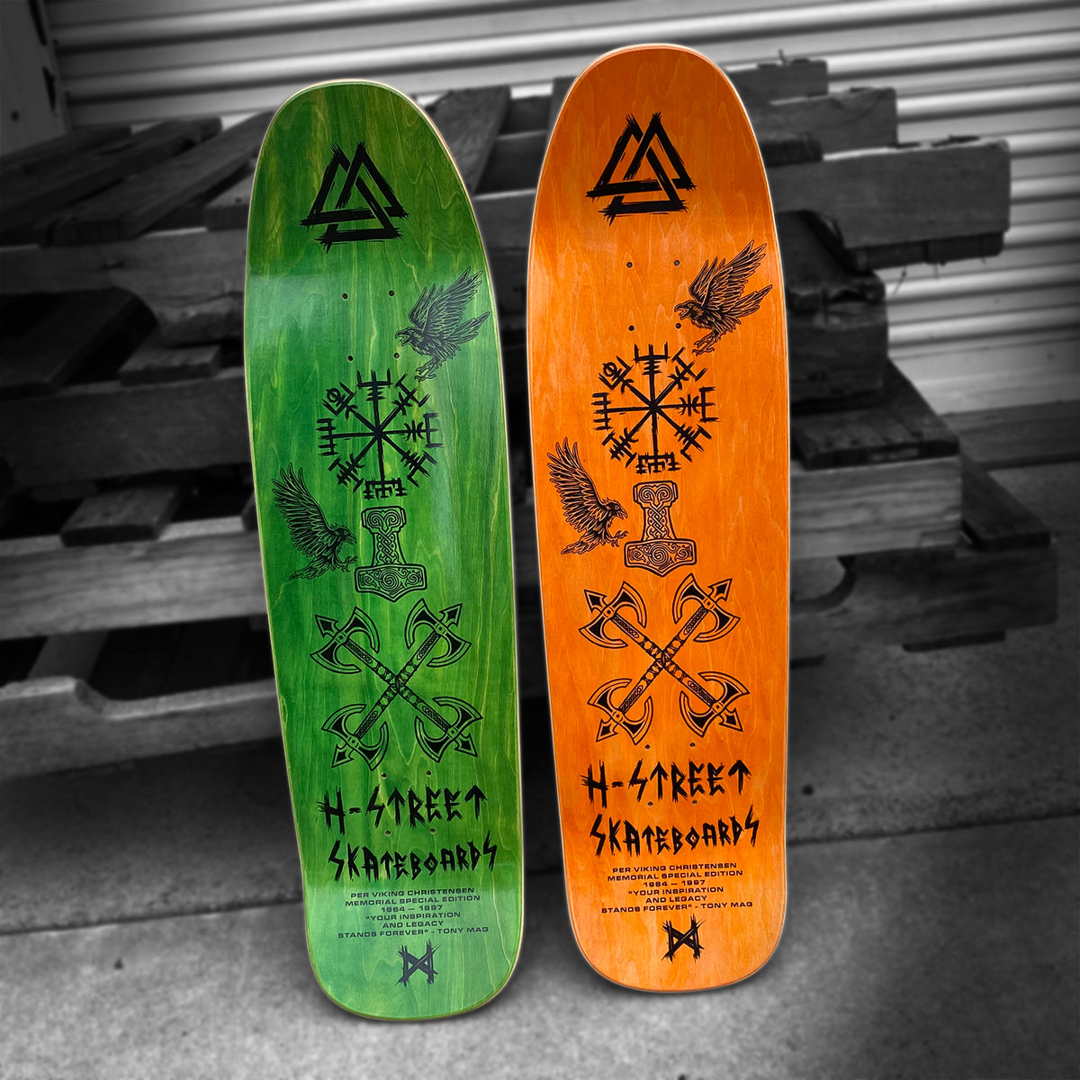 Two PER VIKING MEMORIAL SERIES longboards by Woodchuck stand upright in Vällingby—one green, one orange—featuring black Norse-inspired raven and rune designs, merging skate culture with Viking artistry.