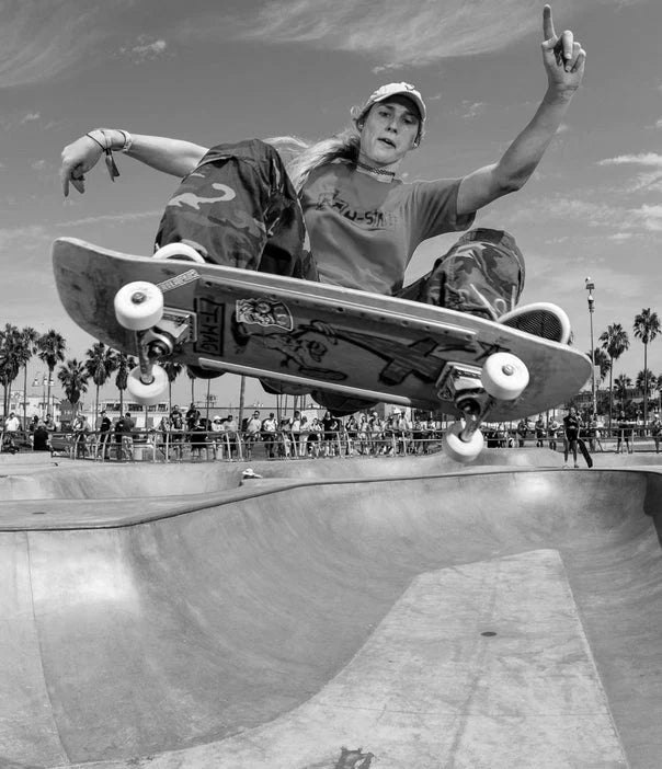 A skateboarder performs an aerial trick above a skatepark bowl, with palm trees and spectators in the background under a partly cloudy sky.
