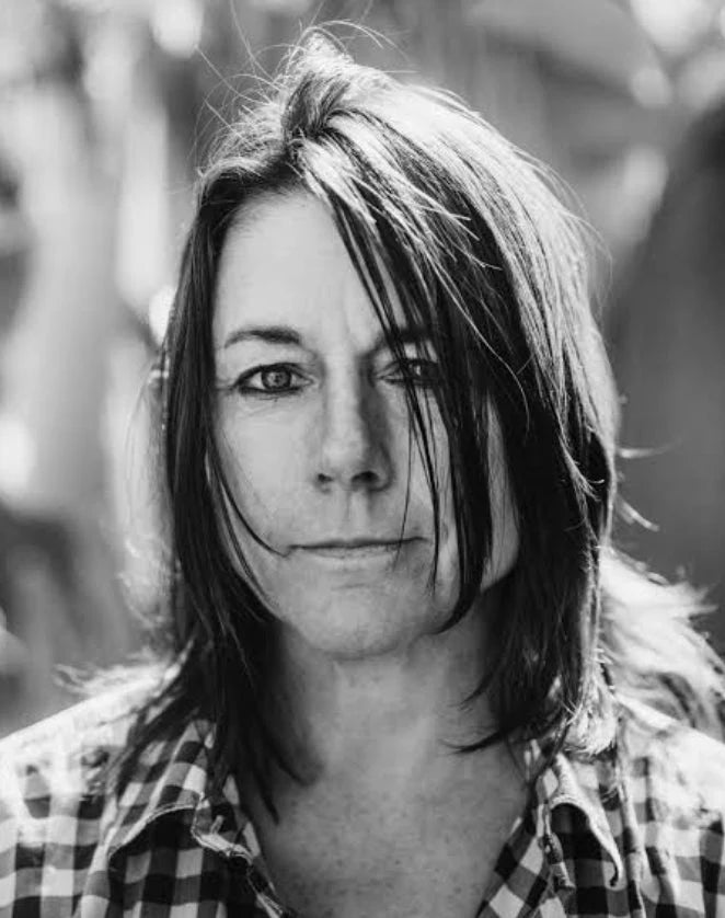 Black and white portrait of a woman with shoulder-length hair wearing a plaid shirt, looking directly at the camera with a neutral expression and strands of hair partially covering her face.