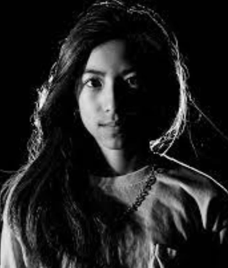 A young woman with long hair is lit dramatically from the side, creating a strong contrast of light and shadow across her face and upper body. The background is completely dark.