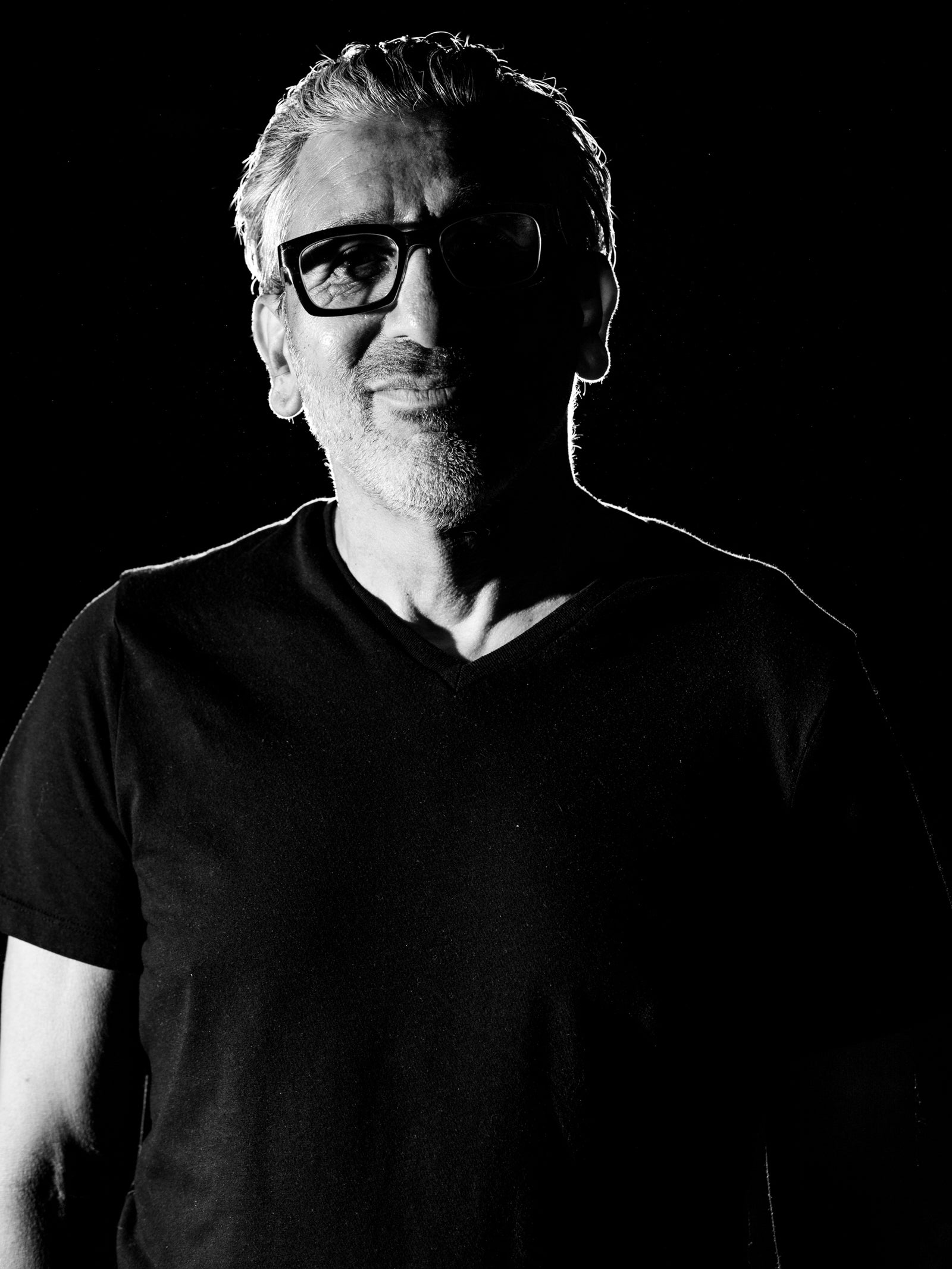 Black and white photo of a middle-aged man with short hair and glasses, wearing a dark V-neck T-shirt, standing against a black background with dramatic lighting highlighting the edges of his face and shoulders.