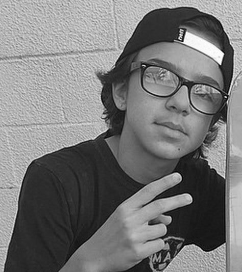 A young person wearing glasses and a backward baseball cap flashes a peace sign with their right hand, standing in front of a textured wall. The image is in black and white.