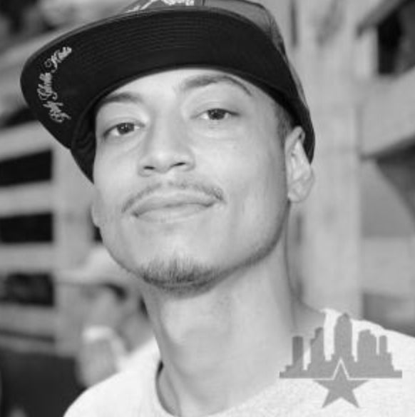 A young man wearing a flat-brimmed cap and a light shirt smiles slightly at the camera. The image is black and white, with a city skyline logo in the bottom right corner.