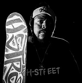 A man wearing a headband holds a skateboard vertically. The image is in black and white with dramatic lighting, highlighting the skateboard’s bold design and the word H-STREET on his shirt.
