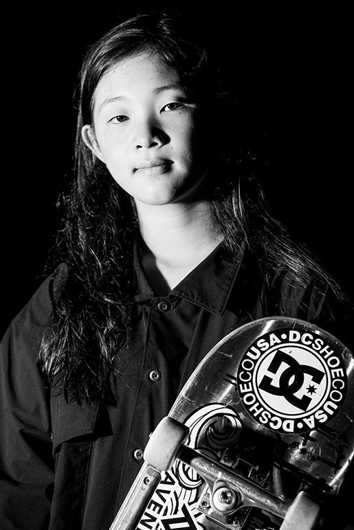 Black and white photo of a young girl with long hair wearing a button-up shirt, holding a skateboard with DC Shoes and Santa Cruz stickers, looking confidently at the camera against a dark background.