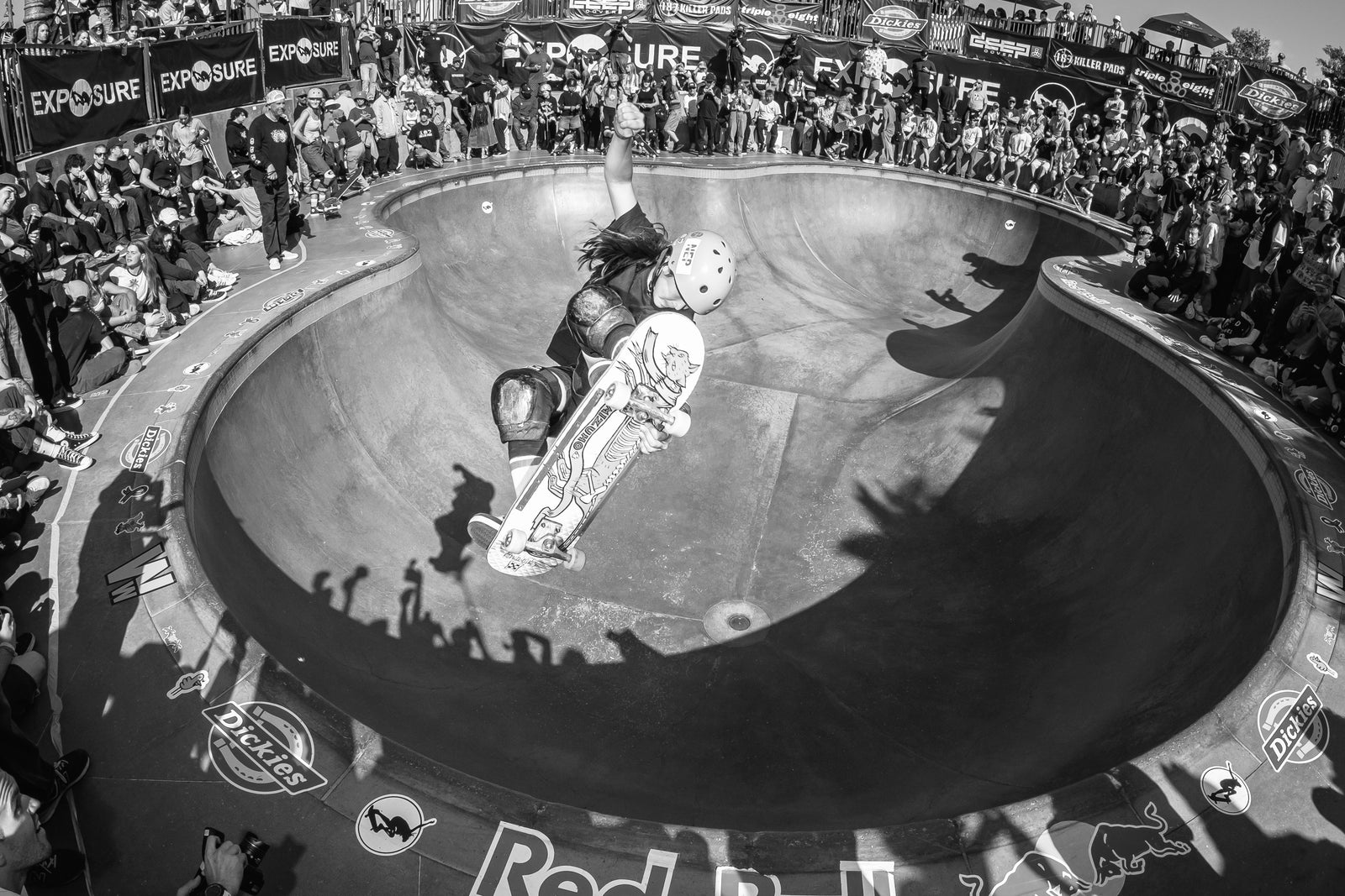 A skateboarder in mid-air performs a trick above a skatepark bowl during a competition, surrounded by a large crowd of spectators and event banners. The image is black and white.