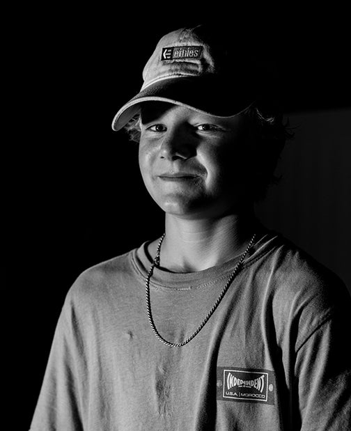 Black and white photo of a young person wearing a cap, a T-shirt, and a chain necklace, looking at the camera with a slight smile. The background is dark and the lighting highlights their face.