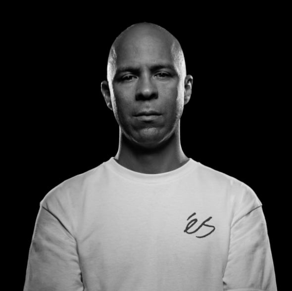 A man with a shaved head wearing a light-colored shirt with a logo, standing against a black background, looking directly at the camera with a neutral expression.