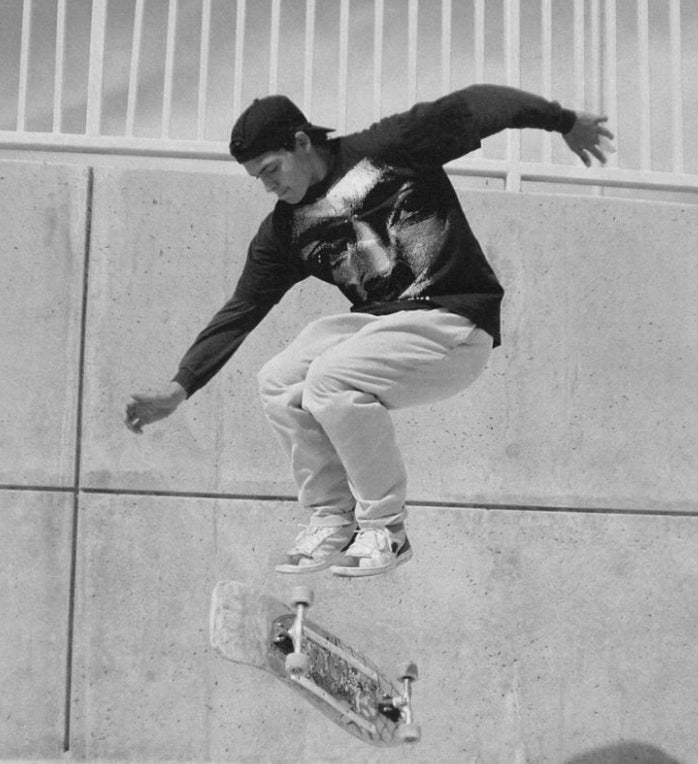 A skateboarder in a backward cap and sweatshirt with a large face print performs a trick mid-air in front of a concrete wall and metal railing.