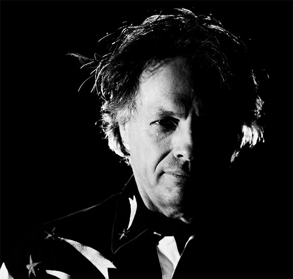 A black-and-white portrait of a middle-aged man with tousled hair, half of his face dramatically lit and the other half in shadow, wearing a shirt with a star pattern.