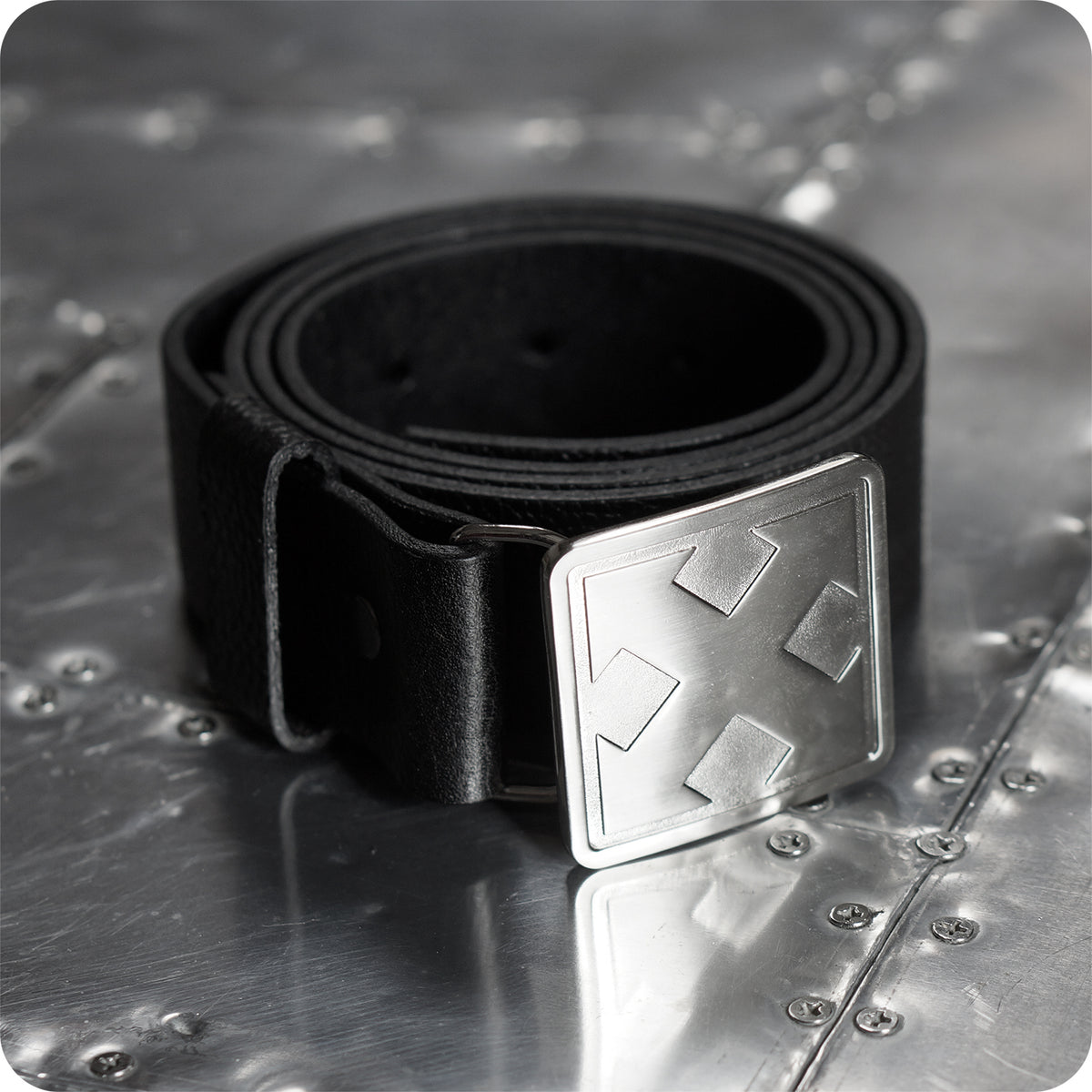 The H-STREET BOMBER BELT by China Factory, a handmade leather belt with a heavy metal buckle featuring a raised cross-arrow design, is rolled and displayed on a riveted metal surface.