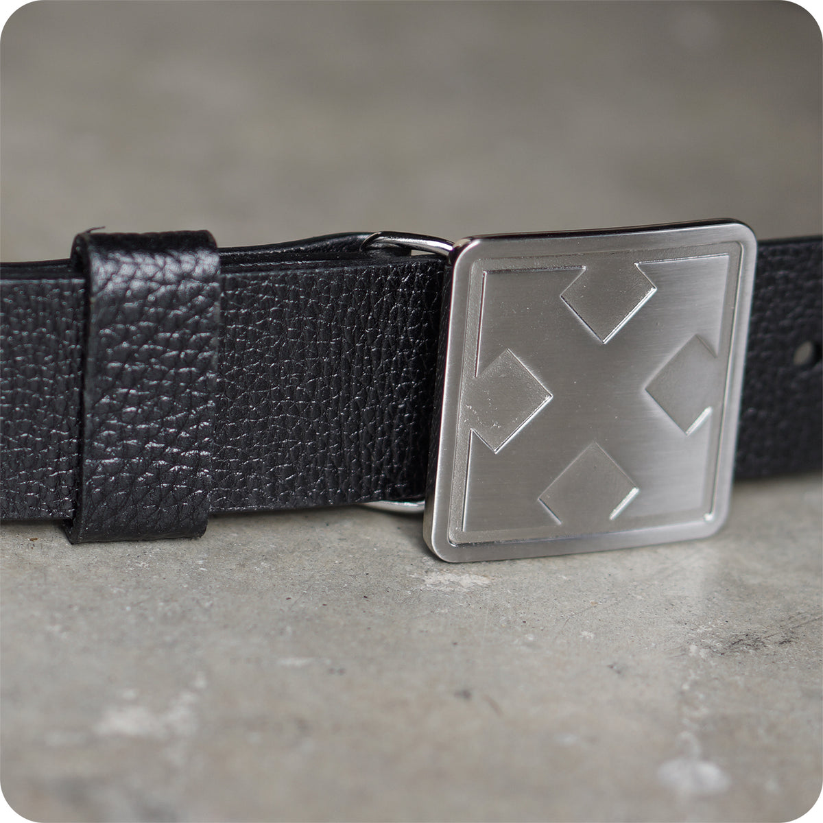 Close-up of the handmade H-STREET BOMBER BELT by China Factory, featuring a shiny metal buckle with a geometric cross and diamond design, displayed on a gray concrete surface.