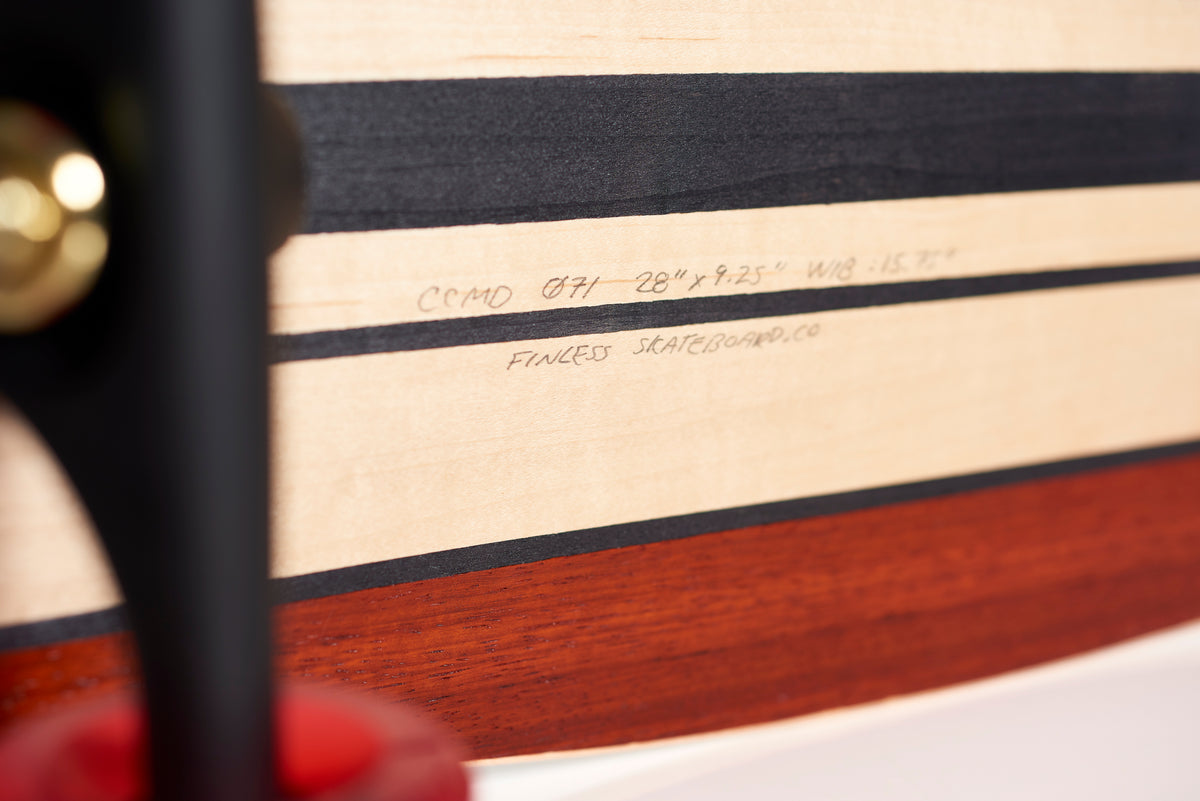 Close-up of a Finless FINLESS COMPLETE longboard deck by Finless, showing handwritten specs on light wood with black and brown stripes; part of the truck and red wheel also visible.