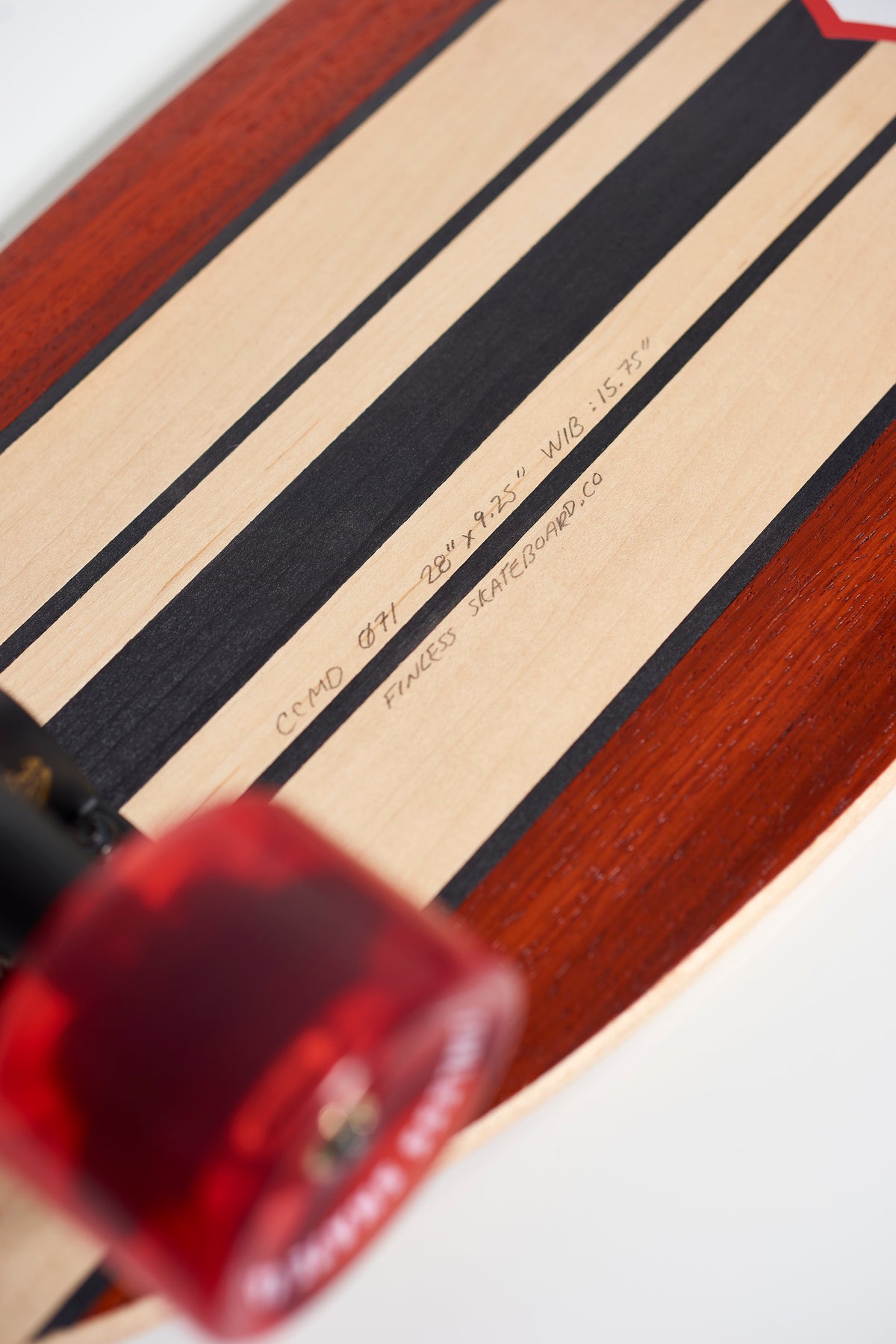 Close-up of the FINLESS COMPLETE by Finless, featuring a handcrafted deck with alternating dark and light wood stripes, red edges, translucent red wheels, and visible specs on its retro surf-style design.
