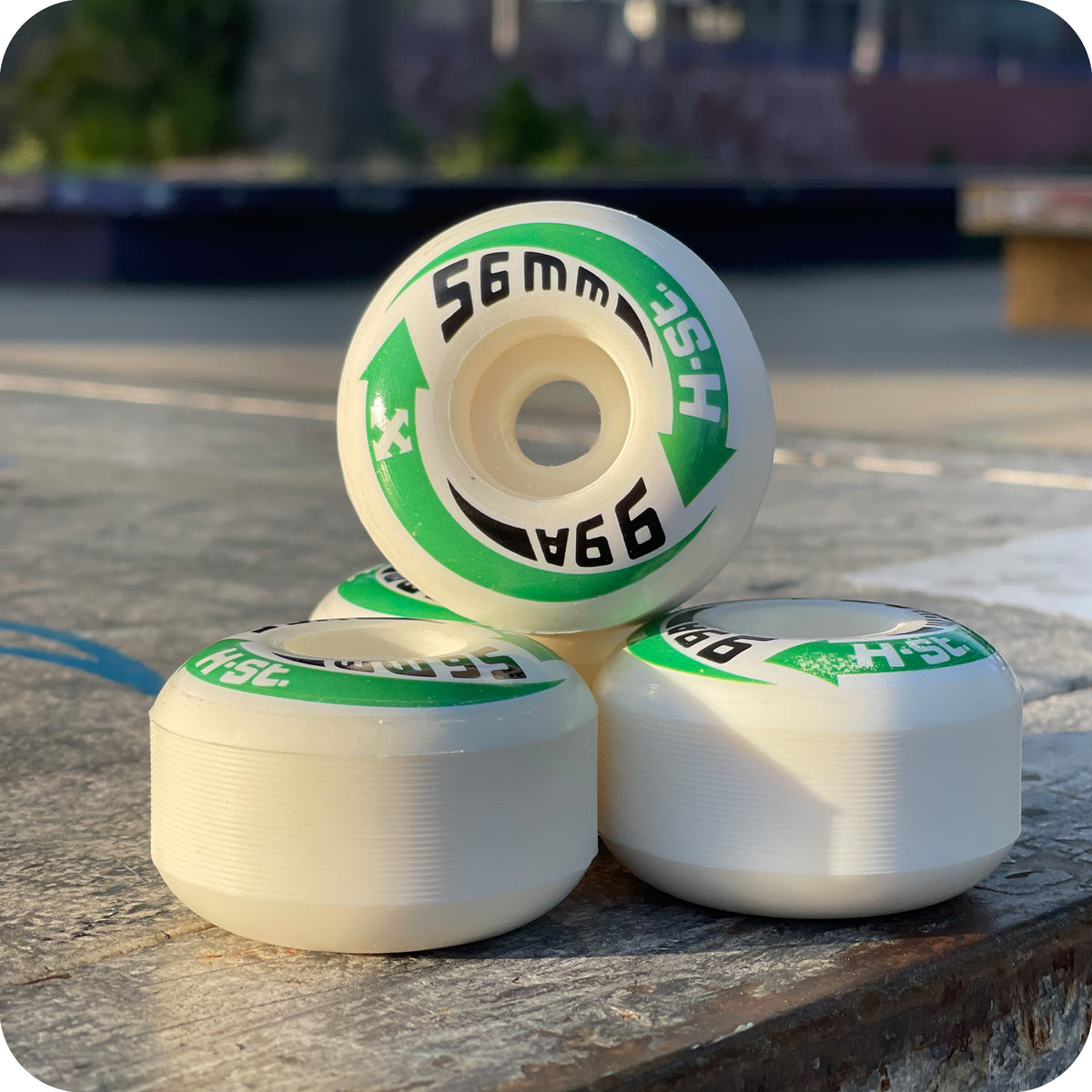 Four white H-STREET LOGO WHEELS by PriPro Polymers, featuring green and black "HSC 56mm 99A" labels, are stacked outdoors on concrete, sunlight highlighting their smooth high rebound urethane.