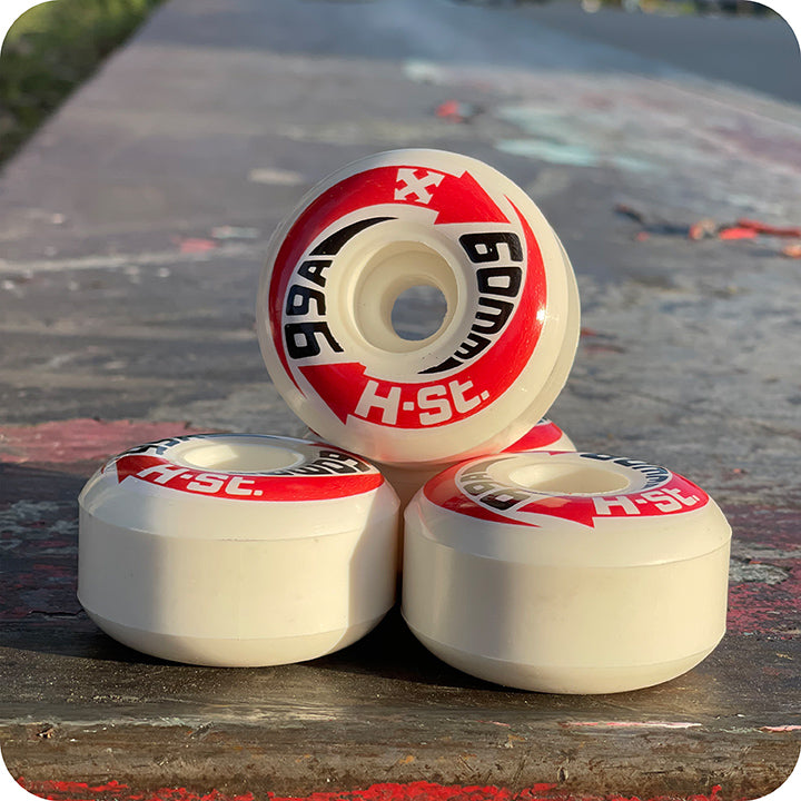 Four white PriPro Polymers H-STREET LOGO WHEELS, “99A 60mm H-ST” with red and black graphics, are stacked outdoors on concrete. Sunlight highlights their clean look and high rebound urethane construction.
