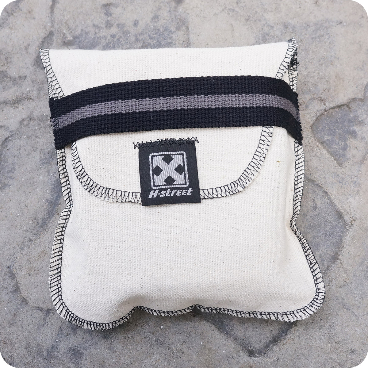 The H-STREET BOMBER BELT by China Factory is a beige fabric pouch with a black and gray striped flap, heavy metal buckle, X logo, and branding on the front, displayed on a stone surface.