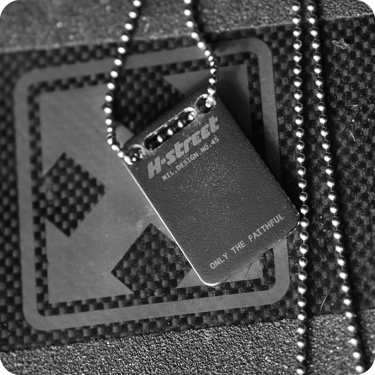 A black and white image shows the H-STREET MARK LOGO DOG TAG by China Factory, a metal skateboarding necklace with ONLY THE FAITHFUL and the Mark Logo engraved on a ball chain, resting on a textured black surface with geometric patterns.