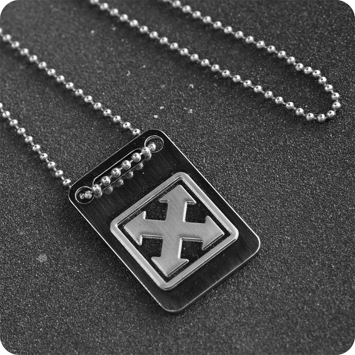 The China Factory H-STREET MARK LOGO DOG TAG necklace features a metallic tag with a raised "X" design on a ball chain, perfect for skateboarding fans, and is shown on a dark, textured surface.