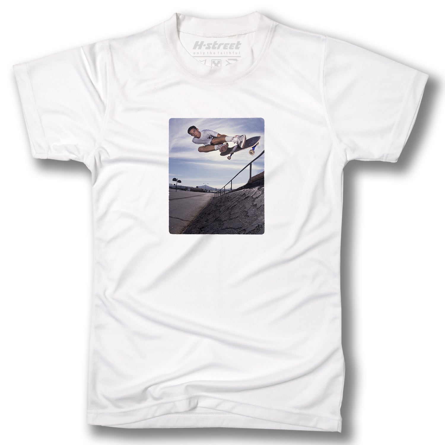 The Minuteman MATT HENSLEY PHOTO TEE is a white skateboarding shirt with a digital print of a skateboarder doing a trick in mid-air above a concrete ledge, set against an urban backdrop and blue sky.
