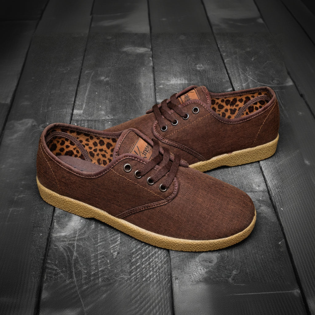 A pair of coffee brown color sneakers with a canvas upper, rubber outsole, and a leopard print insole, positioned on a wooden surface.
