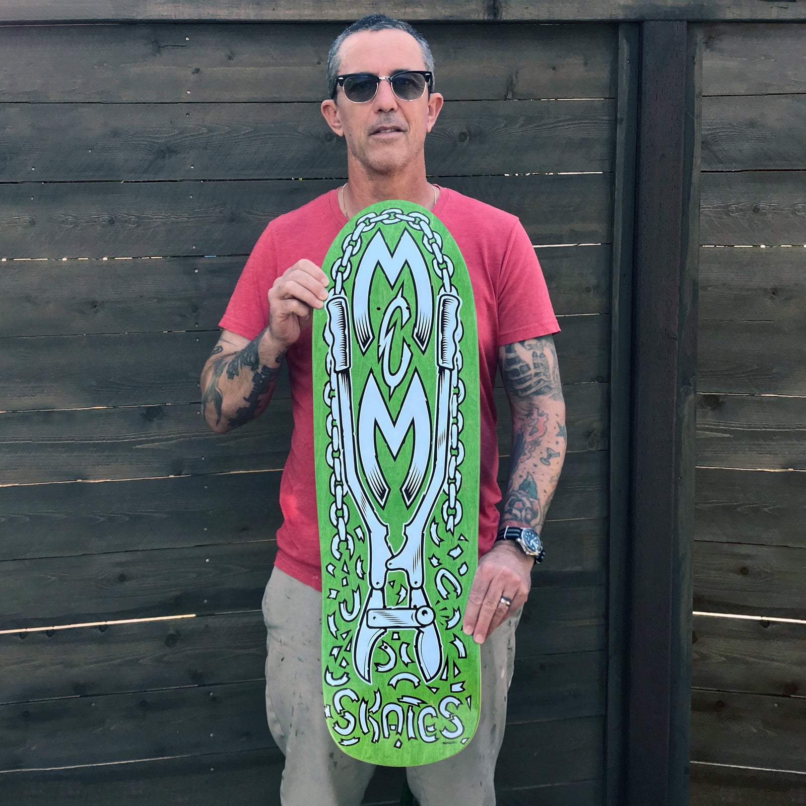 A man in sunglasses and a red t-shirt holds a Street Plant M&M SKATES HENSLEY X VALLELY green skateboard featuring bold chains, lettering, and stylized white and gray art, standing in front of a wooden fence.