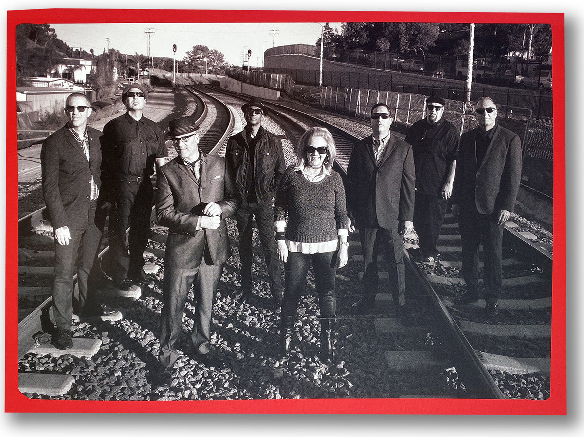 Eight adults in dark clothes and sunglasses pose on railroad tracks, channeling a Spy Kids ska band vibe. The black and white photo with a red border showcases SPY KIDS </p> COVERT ACTION by Matt Hensley. Trees, fences, and signals appear behind them.