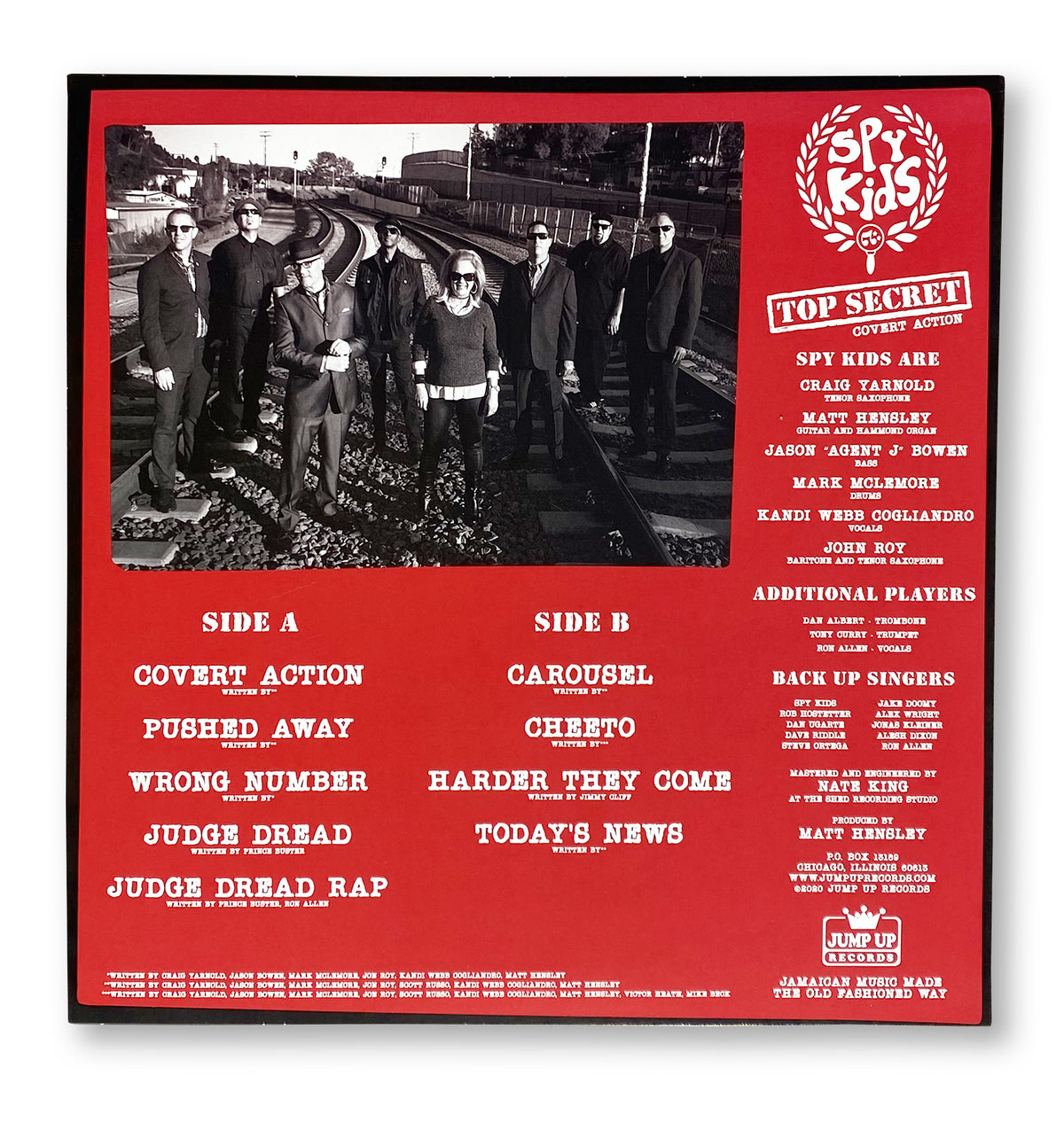 A black-and-white photo shows eight people in suits and sunglasses on train tracks. The red border of SPY KIDS COVERT ACTION by Matt Hensley lists album details, band names, credits, and highlights Jamaican music influences.